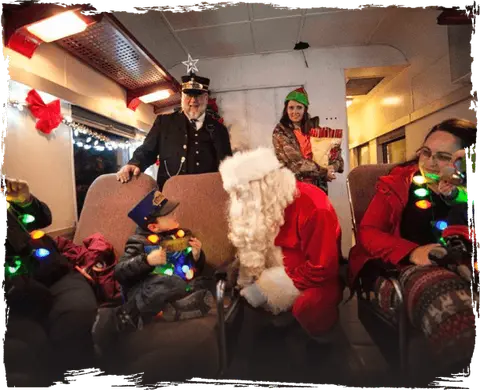 Santa Claus and a train conductor visit passengers inside a decorated train car.