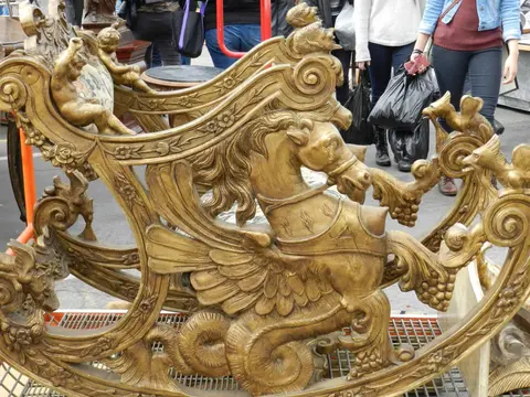 An ornate golden carved wooden piece, possibly a sleigh or carriage, is displayed outdoors near people.