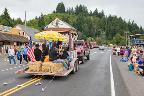A parade float with people sitting on hay bales moves down a street with spectators watching.