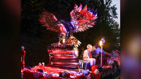 A parade float features a large illuminated eagle perched on a scroll.