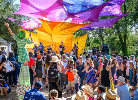 A crowd of people enjoys an outdoor concert under colorful shade sails, with a performer on stilts.