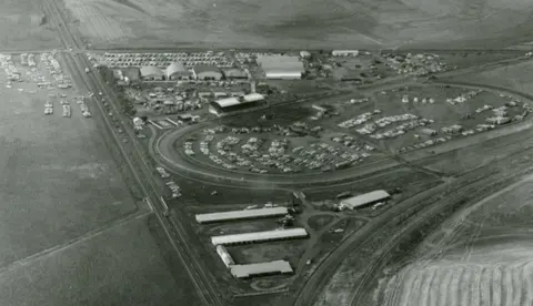 An aerial black-and-white photograph shows a racetrack and surrounding fairgrounds filled with vehicles.