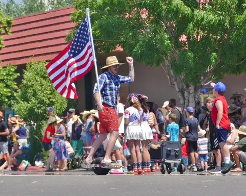 A man rides an electric skateboard while holding an American flag in a parade.