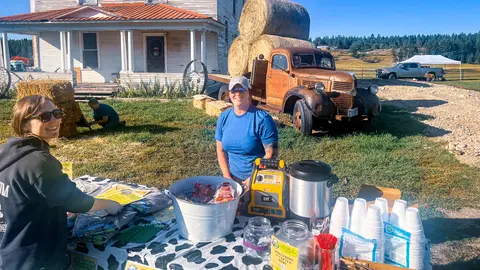 Two women stand behind a table with snacks and drinks outdoors in front of a rustic building and vintage truck.