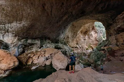 A hiker stands inside a large natural rock cave with an arched opening.