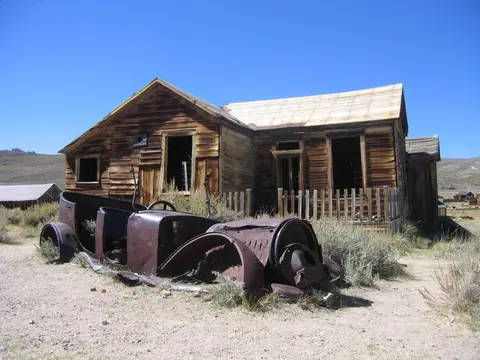 An old wooden building stands behind the rusted frame of an antique car.