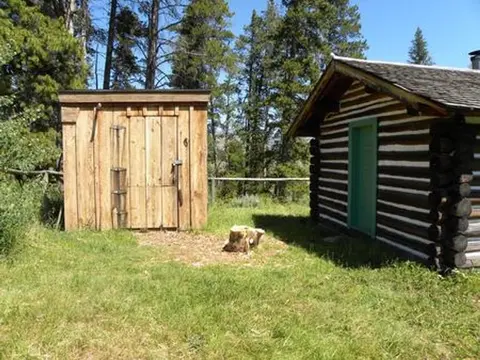 A log cabin and a wooden shed stand in a grassy clearing with trees.