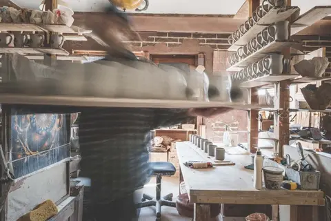 A person working inside a pottery workshop carrying a board of ceramic items.