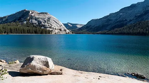 A scenic view of a calm alpine lake surrounded by mountains and forests under a clear blue sky.