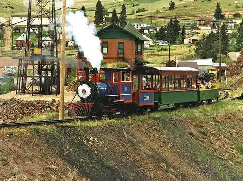A small steam train travels on tracks through a hilly landscape near buildings and a wooden tower.