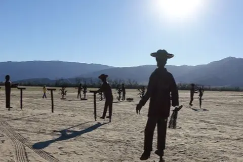 Metal sculptures of figures stand in a desert landscape against mountains.
