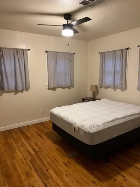 A bedroom with a bed, hardwood floors, and windows with curtains.