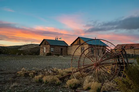 Old rustic buildings and farm equipment sit under a colorful sunset sky.