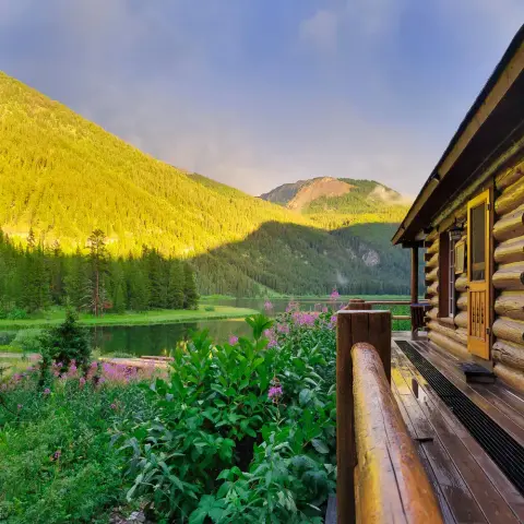 A log cabin porch overlooks a serene lake and forested mountain landscape.
