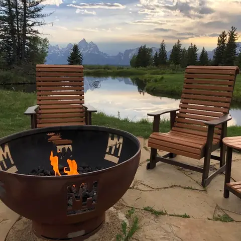 Two wooden chairs sit near a fire pit overlooking a body of water and mountains.