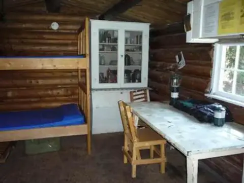 An interior view of a rustic log cabin showing bunk beds, a cabinet, a table, and chairs.