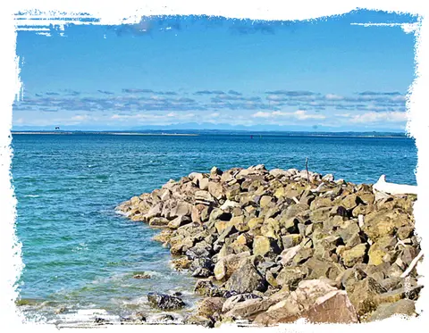 A rocky breakwater extends into a calm blue sea under a cloudy sky.