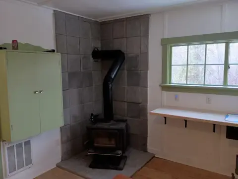A wood-burning stove is installed in the corner of a room with a green cabinet and a window.