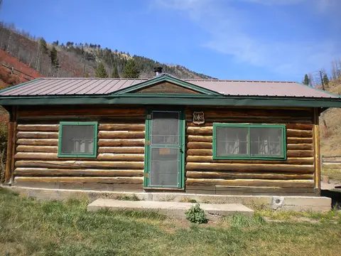 A log cabin sits in a mountain landscape under a clear blue sky.