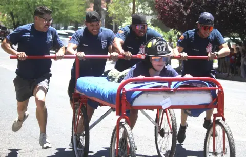 Four firefighters push a bed on wheels with a teammate riding it on a street.