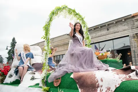 A young woman wearing a tiara and sash waves from a parade float.