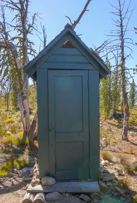 A green outhouse stands outdoors among trees and sparse vegetation under a blue sky.