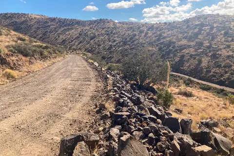 A dirt road winds through arid, hilly terrain under a blue sky with scattered clouds.