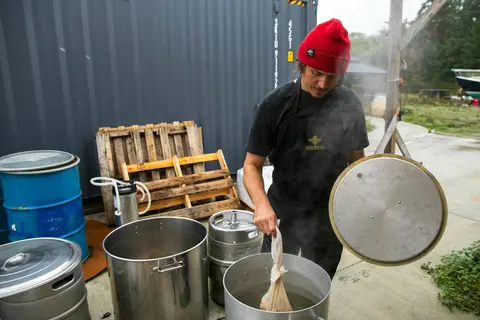 A person prepares a brew outdoors next to large metal pots and containers.