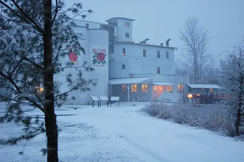 Exterior view of a large white mill building during a snowfall.