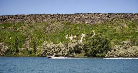 A motorboat speeding across a river with waterfalls and a rocky cliff in the background.