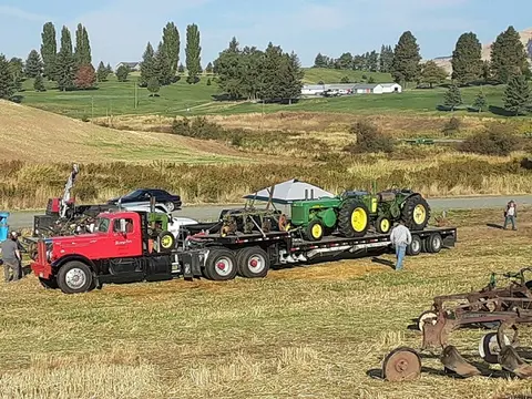 A red semi truck carrying vintage tractors on a flatbed trailer in a grassy field.