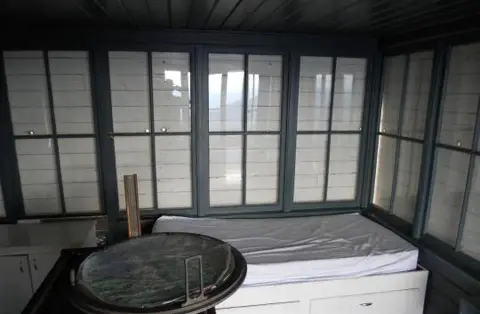 An interior view of a fire lookout cabin with a bed and windows.