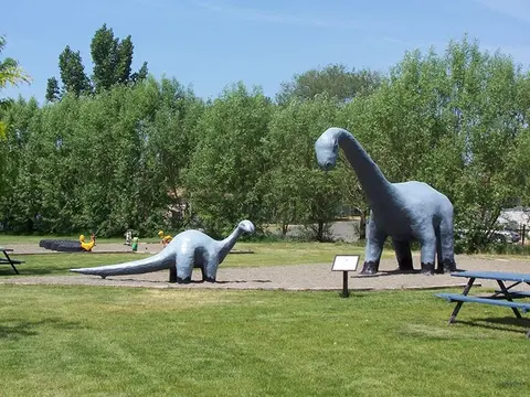 Two dinosaur statues stand in a grassy park near picnic tables.