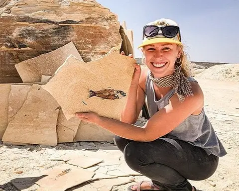 A smiling woman holds a slab of rock containing a fossilized fish outdoors.