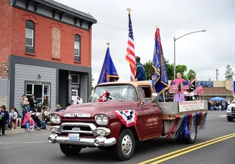 A decorated vintage pickup truck carries flags and people in a parade on a city street.