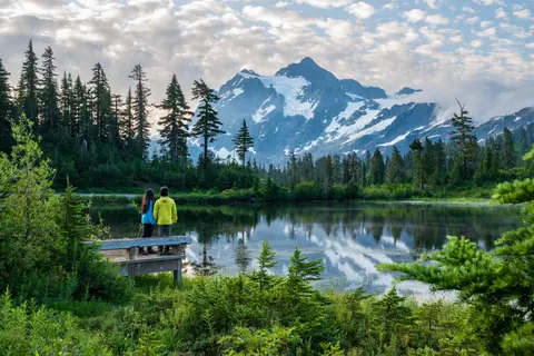 Two people stand on a wooden viewing platform looking out at a lake reflecting a snow-capped mountain range and evergreen forest.
