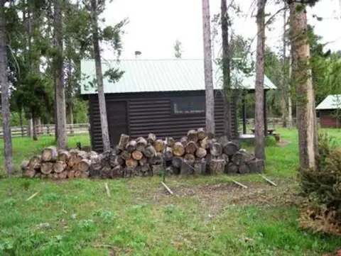 A rustic log cabin sits among trees with a pile of split firewood in the foreground.