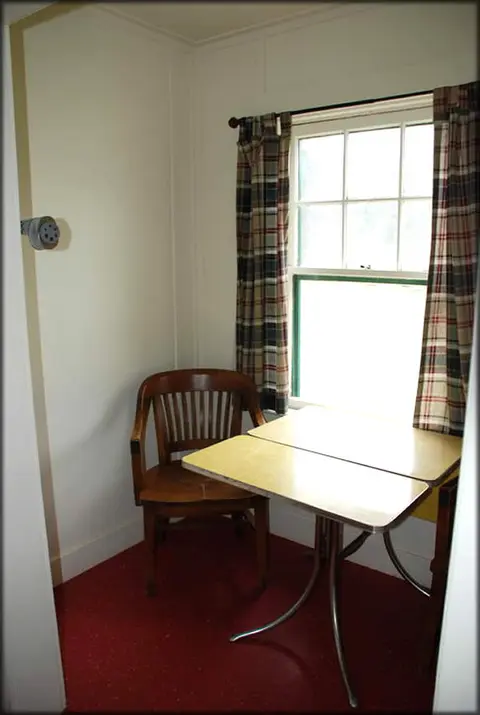 An interior corner with a small table, a wooden chair, and a window with plaid curtains.