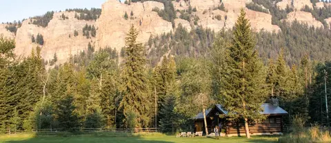 A rustic log cabin sits in a clearing surrounded by tall pine trees and towering rocky cliffs.
