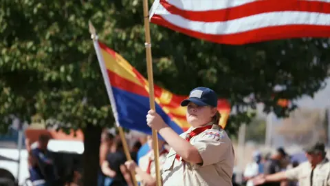 A young scout carries flags in a parade outdoors.