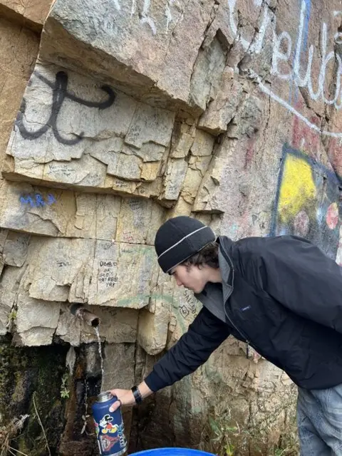 A young man fills a water bottle from a pipe flowing out of a rocky cliff.