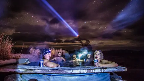 A group of people enjoy a hot tub under a starry night sky with a bright light beam.