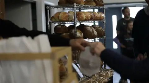 People buying bread at a bakery counter with a rack of fresh bread in the background.