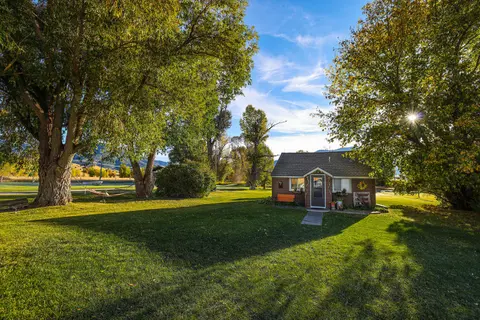 A small cabin sits on a green lawn surrounded by large trees under a blue sky.