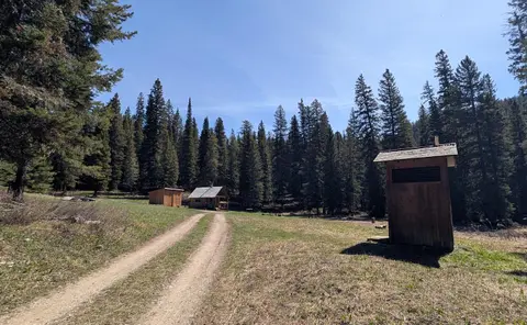 A dirt road leads past a small outhouse toward a cabin in a forest clearing.