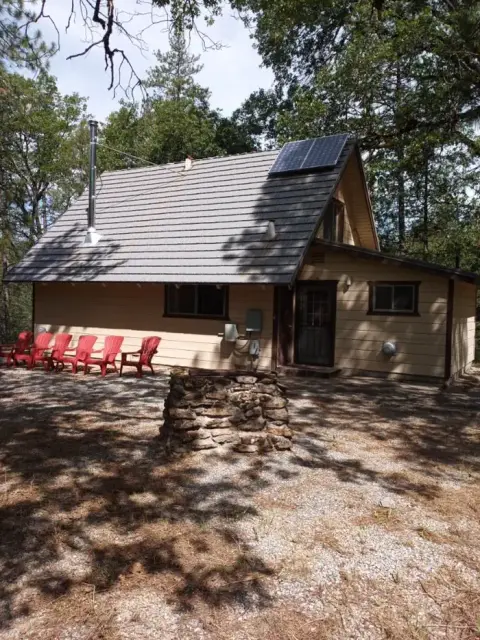 An exterior view of a cabin in the woods with red chairs and a stone fire pit.