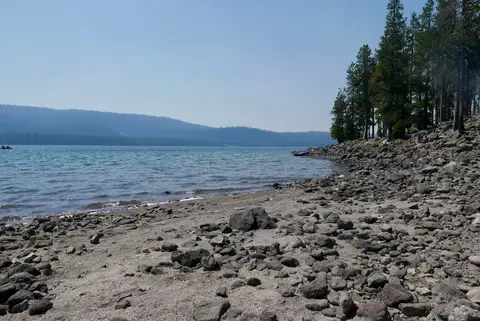 A rocky shore beside a lake with trees and distant mountains.
