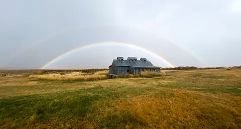 A double rainbow arches over an old rustic building in a grassy field.