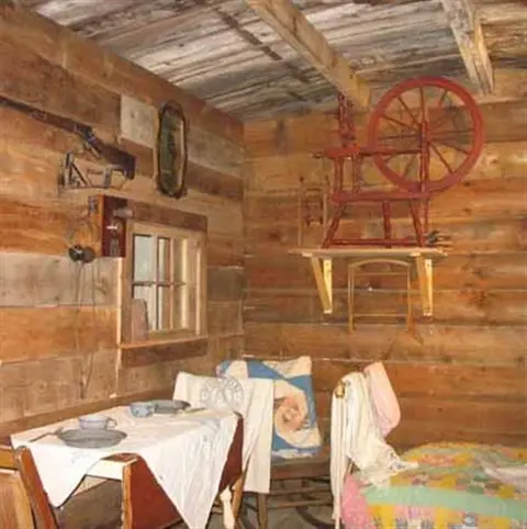 The interior of a rustic log cabin shows historic household items, a dining table, and a bed.
