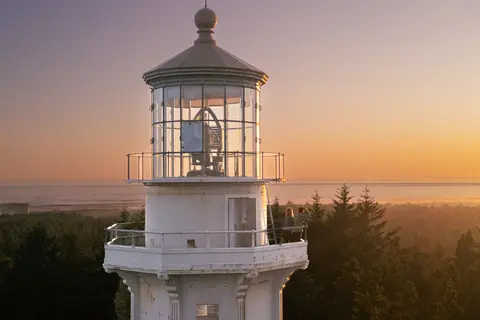 A lighthouse tower stands outdoors at sunset with people standing on the balcony.